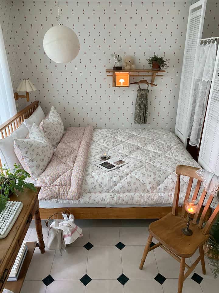 Cozy bedroom with white floral patterned wallpaper, wooden frame bed with quilted duvet, and vintage-style chair