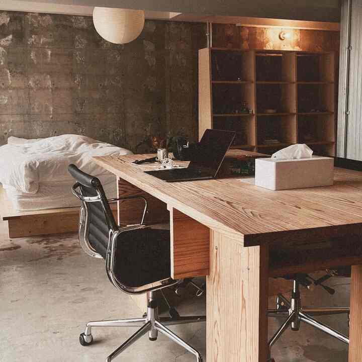 Modern studio apartment with brown wooden desk, black office chair, bed, and industrial style concrete walls