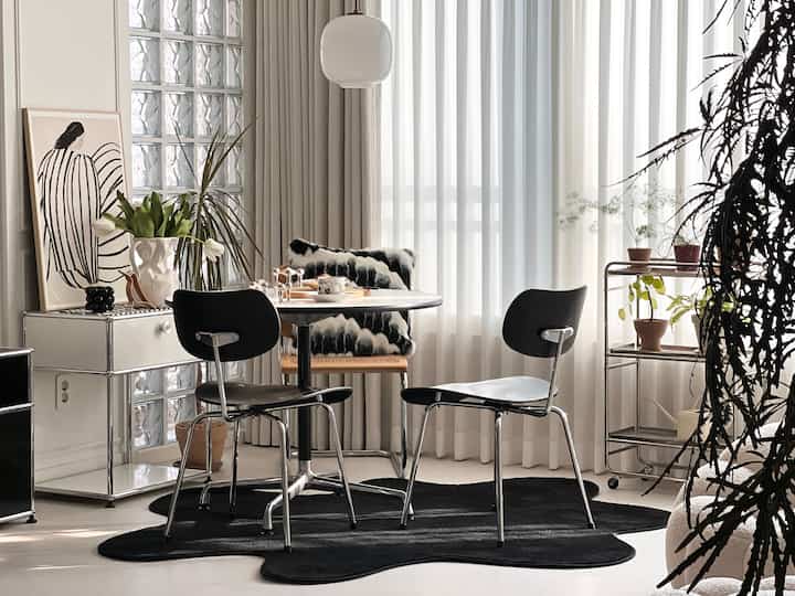 白黒トーンのリビング、ミッドセンチュリーモダンスタイルの丸テーブルと椅子、大きなカーテンと植物が配置された雰囲気のあるインテリア