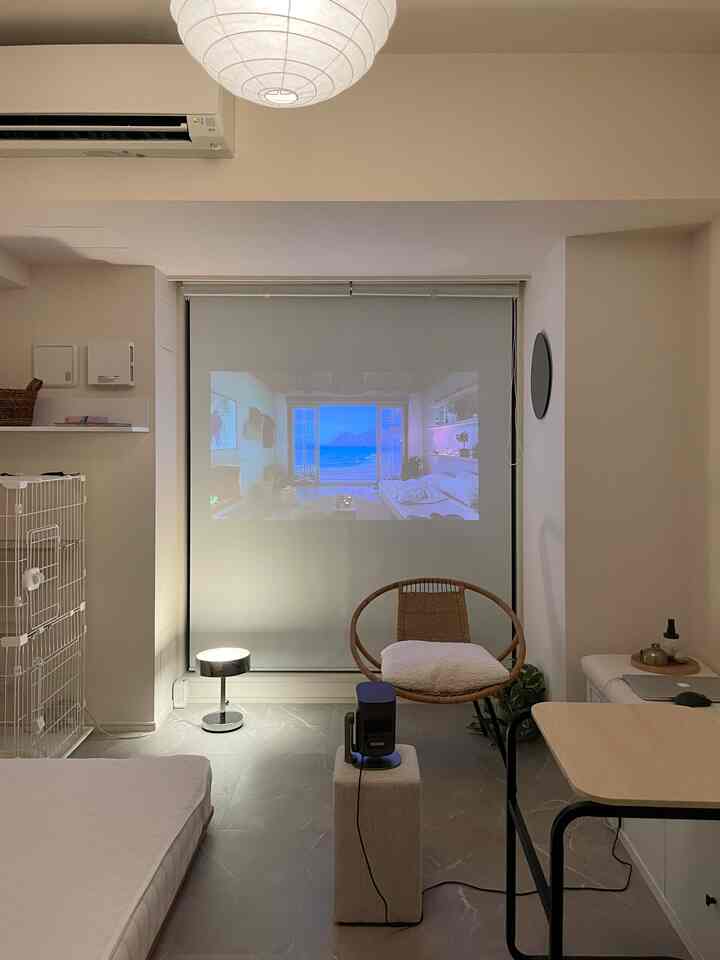 白とベージュのトーンでまとめられた小さなワンルーム、ラタンチェアとプロジェクタースクリーンが配置されたミニマルなホームシネマ空間