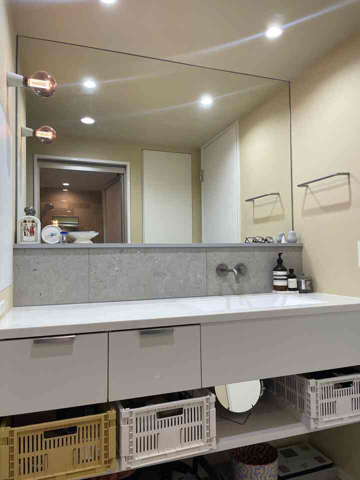 ベージュの壁と白い洗面台が配置された浴室、壁付け照明と大きな鏡が調和するシンプルで実用的なインテリア