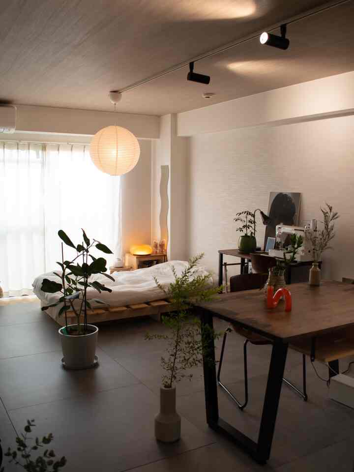 A natural modern studio apartment bedroom with soft wood tones and white walls, featuring a dining table and abundant plants