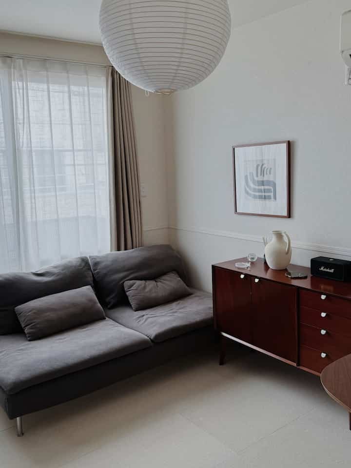 淡いベージュの壁とダークグレーのソファ、ブラウンのキャビネットが配されたシンプルな北欧風リビング、ペンダントライト付き