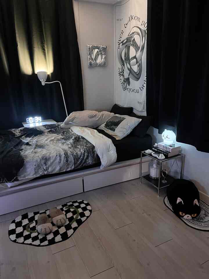 黒と白のトーンが特徴のワンルーム寝室で、ベッド、ファブリックポスター、ムード照明、カーペットが調和する雰囲気のあるインテリア