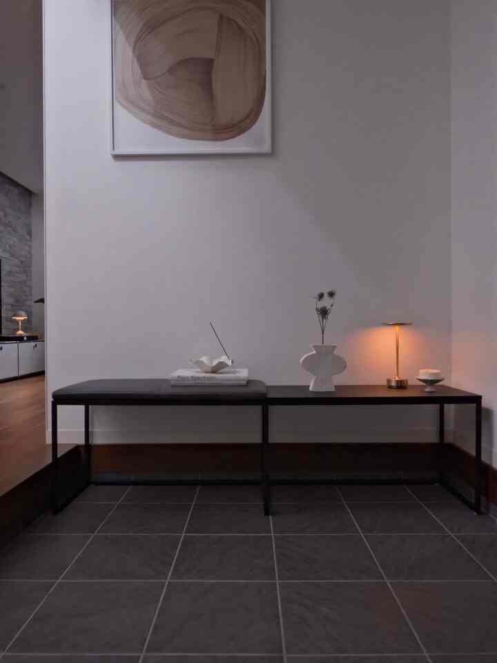 グレーのタイルと黒いベンチが調和する玄関、テーブルランプと抽象ポスターが配置されたミニマルな空間