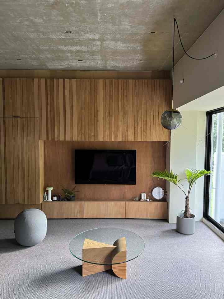 ナチュラルな木目調の壁とグレーのカーペットが調和するモダンなリビング、ガラスのコーヒーテーブルとペンダントライトが配置された印象的な空間