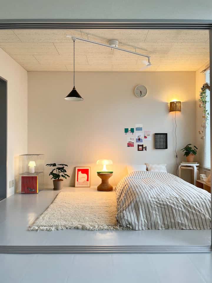 白を基調とした寝室、ブラウンのアクセントが効いた空間にヴィンテージのペンダントライトと自然素材のスツールが配置された雰囲気のあるインテリア