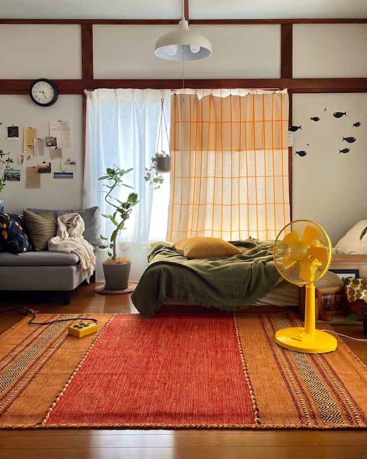 オレンジと黄色のカーテンとラグ、グレーのソファとベッドが配置されたナチュラルな雰囲気のワンルームのリビング兼寝室
