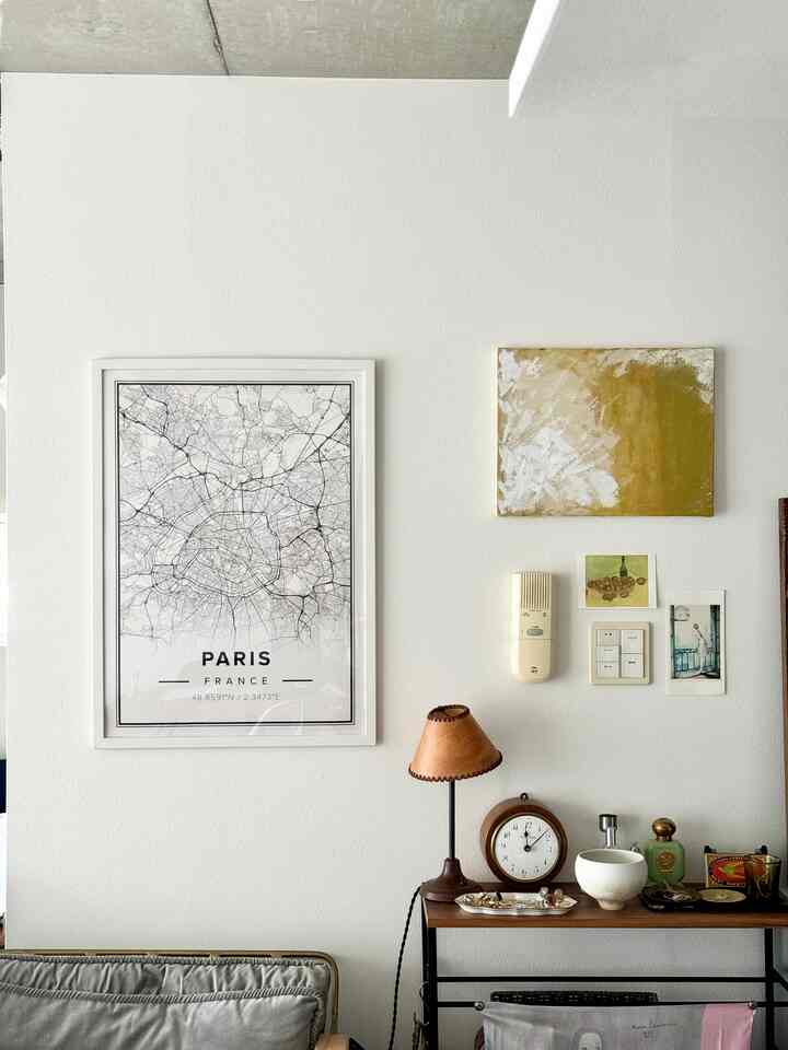 白い背景のリビング、パリ地図と抽象画の額縁、木製の棚にヴィンテージ風のテーブルランプと時計が配置された雰囲気のある空間