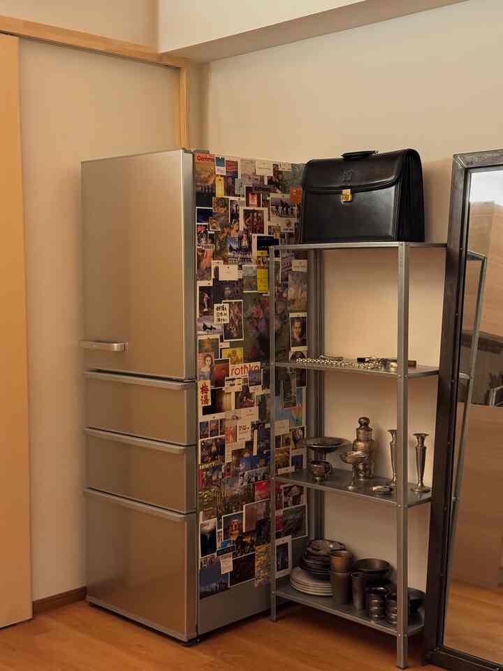 A studio apartment kitchen with beige-toned walls and wood flooring featuring a refrigerator and simple metal shelf in a tidy design