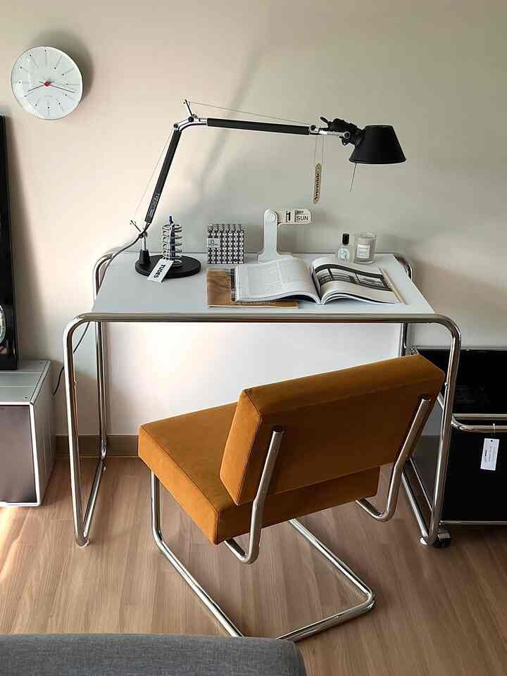 ホワイトの壁と明るいブラウンの床の狭いミニマルなホームオフィス、白いデスクとブラウンのアームチェア、黒いテーブルランプを備えた空間