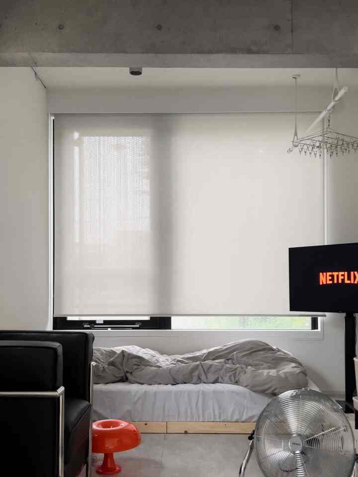 Modern studio bedroom in gray and black tones featuring a bed, armchair, fan, and TV, with natural light filtering through window blinds