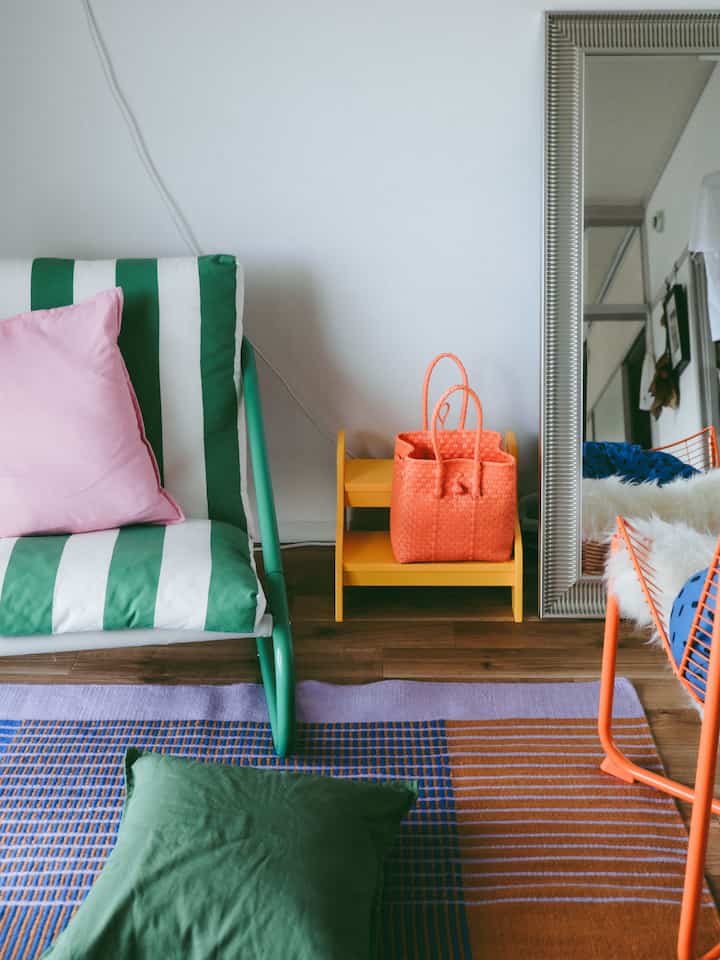 Bright space with green and white striped armchair with pink pillow, orange bag on yellow stool, and a large silver-framed mirror