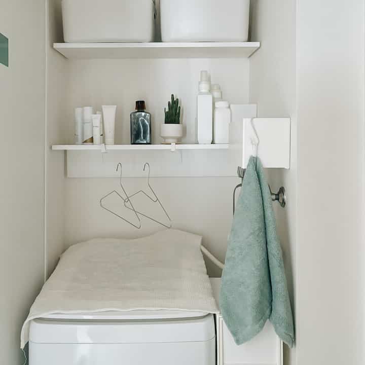 White-toned small laundry room featuring clean storage shelves, washing machine covered neatly, and pale green towels, creating a simple atmosphere