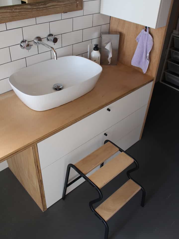 Natural-colored bathroom featuring a white sink, tiled wall, and wooden stool creating a clean atmosphere