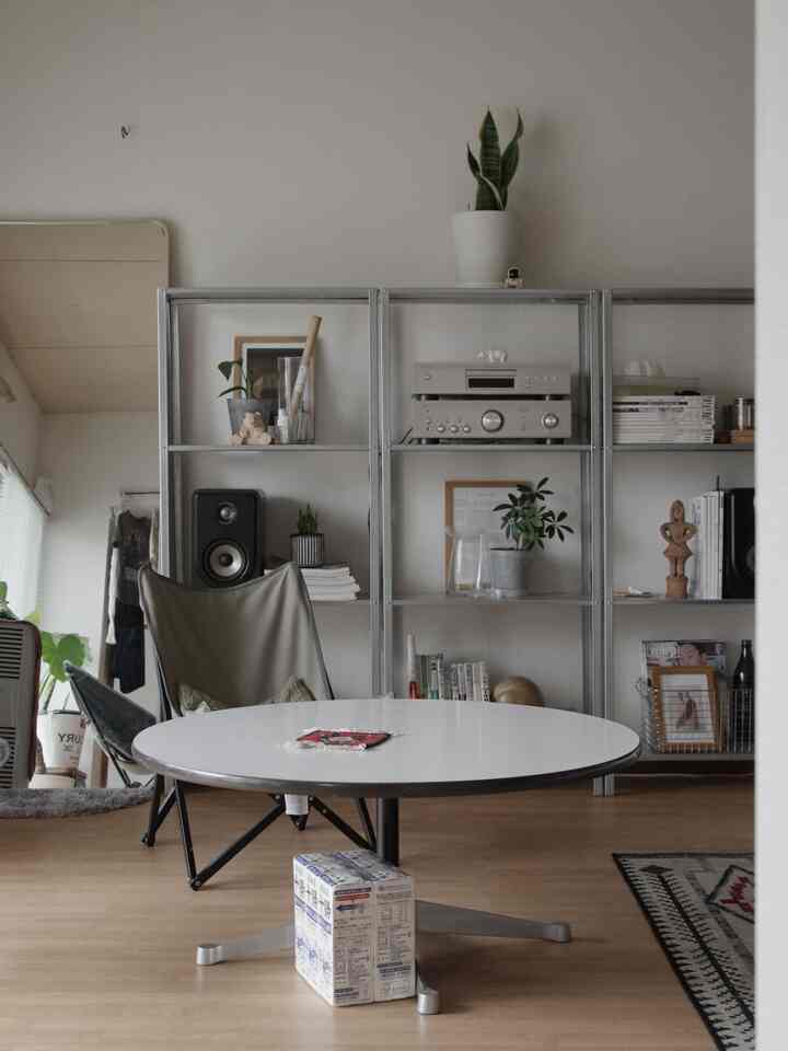 白い壁と茶色い床のモダンなリビング、中央に丸いテーブルとベージュのアウトドアチェアが配置された印象的な空間