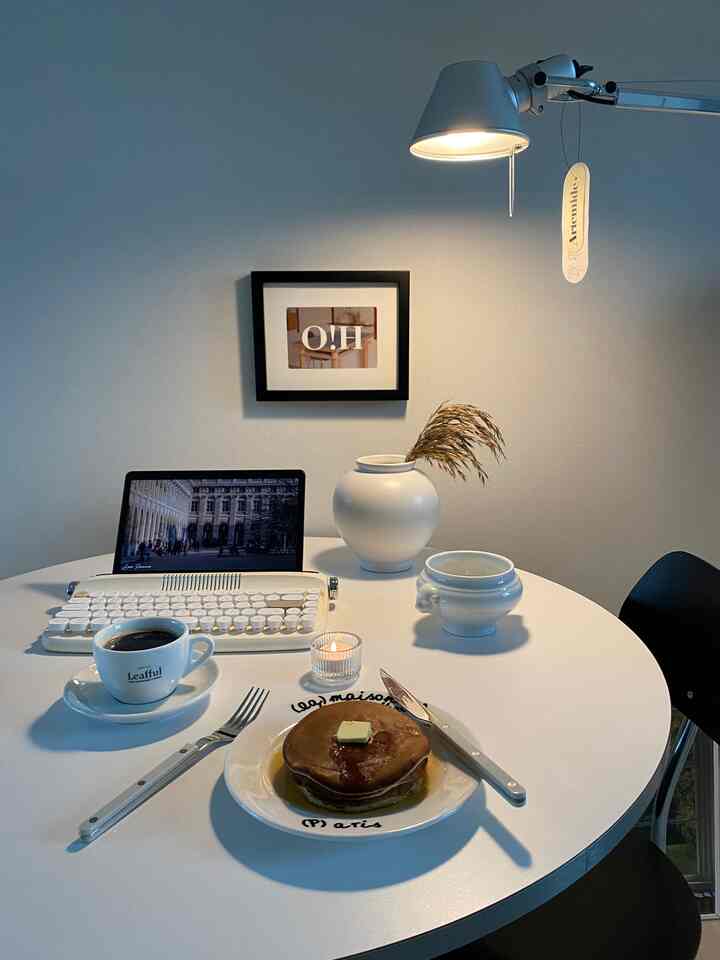 白いトーンのダイニングルーム、丸いテーブルにホームカフェの雰囲気のパンケーキとコーヒー、作業用機器が配置されたシンプルな空間