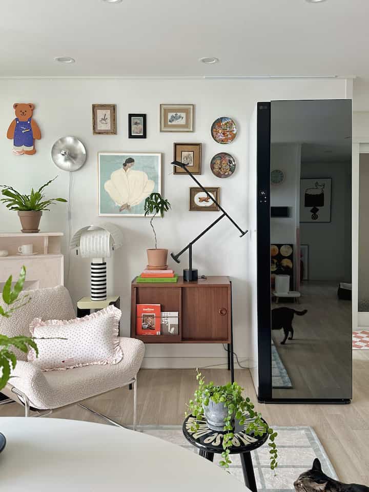 A white and brown toned living room featuring a nightstand, stool, wall sconce, multiple artworks, and plants creating a cozy space