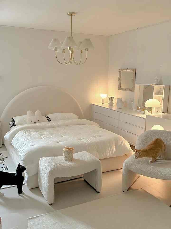 Ivory and white toned bedroom featuring rounded headboard bed, soft armchair, and two cats in a cozy atmosphere