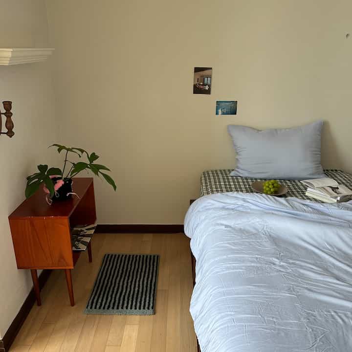 Natural modern bedroom with white walls and wood floor, featuring a bed with blue bedding and cushion, wooden shelf with a plant, creating a simple and cozy atmosphere