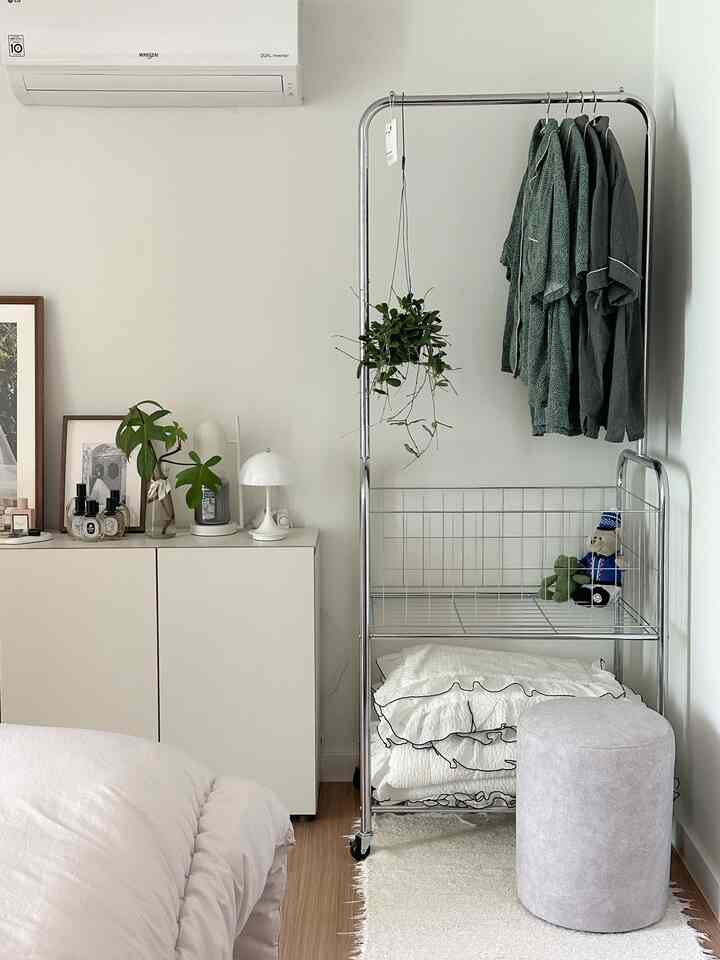 ホワイトとウッドトーンが調和する寝室、移動式スチールハンガーにパジャマが掛けられ、収納された寝具と小さなスツールが配置された雰囲気のあるインテリア