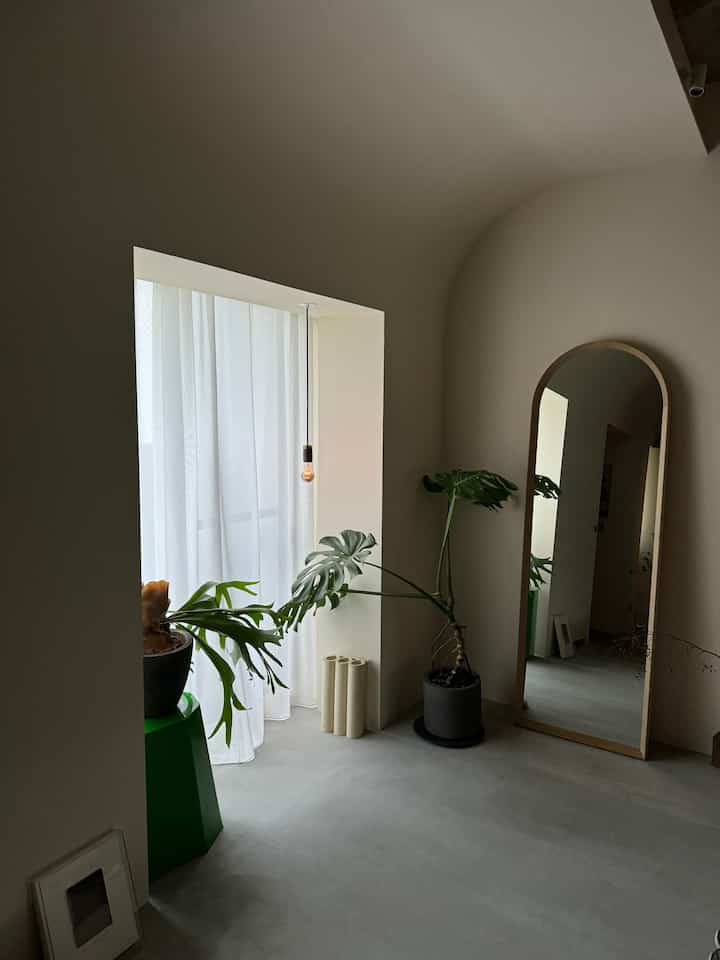 柔らかな曲線の壁と白いカーテンがあるモダンな玄関空間、大きな植物と全身鏡が配置された自然な雰囲気のインテリア