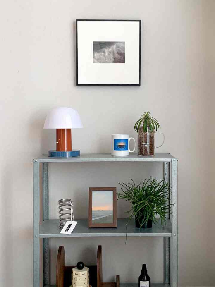 ホワイトとグレーのトーンのモダンな空間、イケアの亜鉛メッキ棚、テーブルランプ、額縁、植物が調和したすっきりとしたインテリア