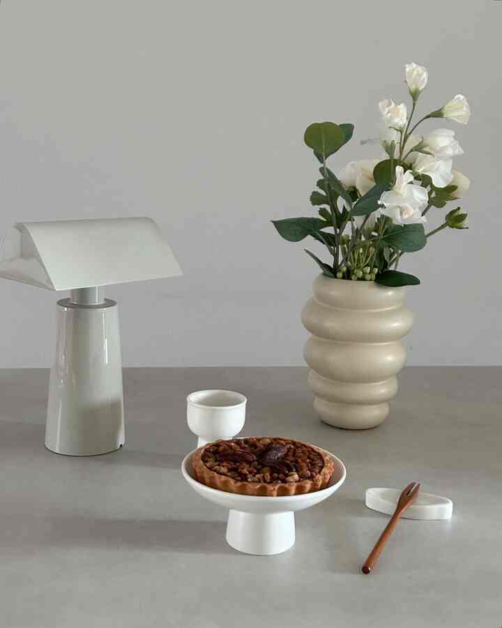 White and beige toned kitchen table featuring a table lamp, vase with flowers, and pecan pie on a simple modern styled setting