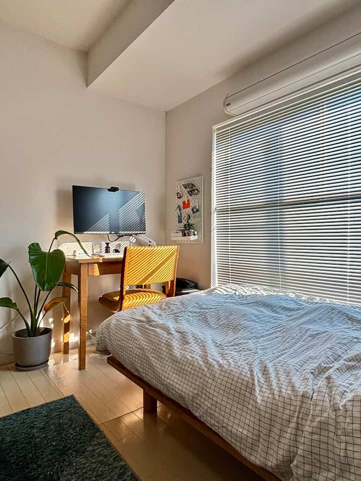 暖かい木目調とナチュラルカラーが調和した一人暮らしの寝室、窓のブラインドとパソコンデスクが配置された居心地の良い空間