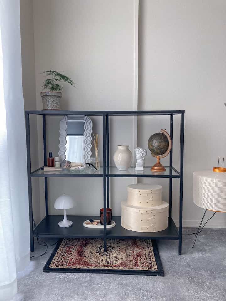 白い壁と明るい床が調和するリビング、黒のメタル棚に鏡や装飾品が配置されたモノトーンのインテリア