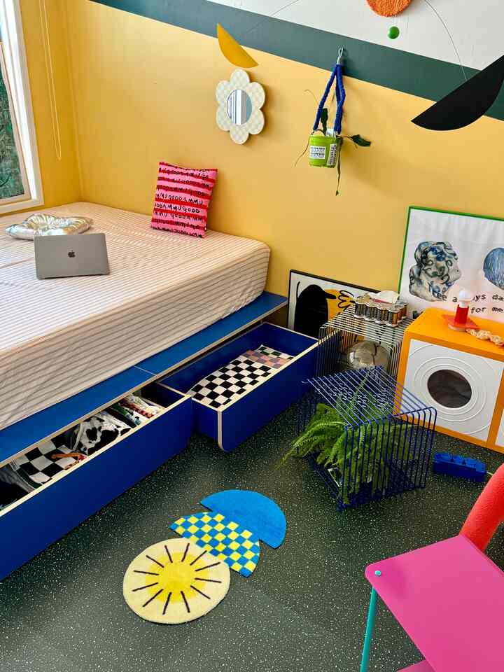 A bright and lively bedroom featuring yellow walls, blue storage drawers under the bed, colorful accessories, and Mid-Century Modern style furniture