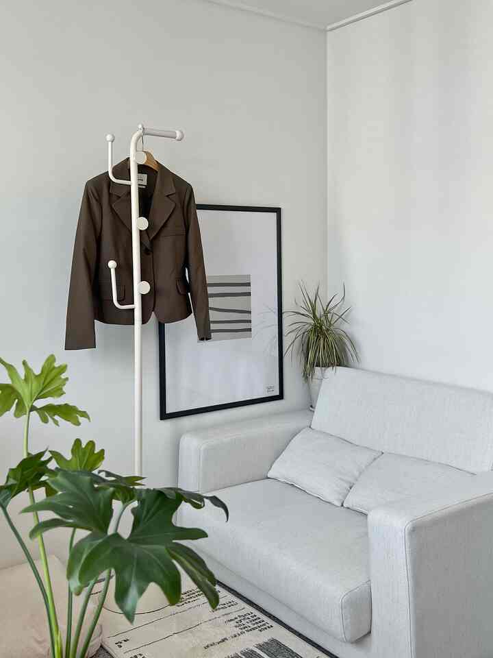 A white and gray toned living room featuring a two-seater sofa, coat rack, and plants in a simple modern setting