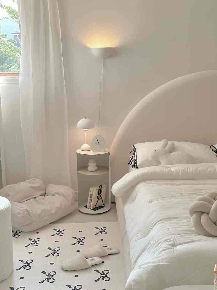 白とベージュのトーンの寝室、丸みを帯びたベッドフレームとシンプルなナイトテーブル、柔らかなラグとクッションが雰囲気のあるインテリア