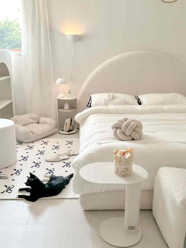 暖かいホワイトトーンの寝室、柔らかな曲線のベッドとホワイトのナイトテーブル、床に横たわる黒い猫がいる雰囲気のあるインテリア