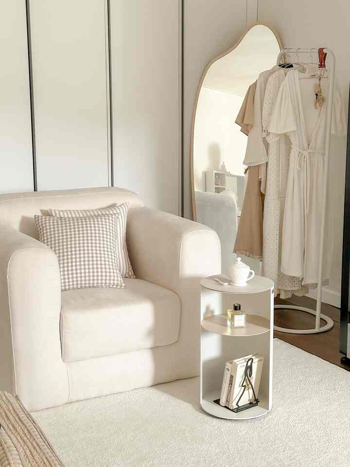 明るいベージュと白のトーンのリビングルーム、ソファと小さなサイドテーブル、全身鏡と洋服ラックが配置されたミニマルで落ち着いた空間