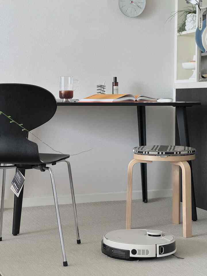 Beige carpeted living room with modern dining chairs, stool, and a white robot vacuum cleaner on the floor