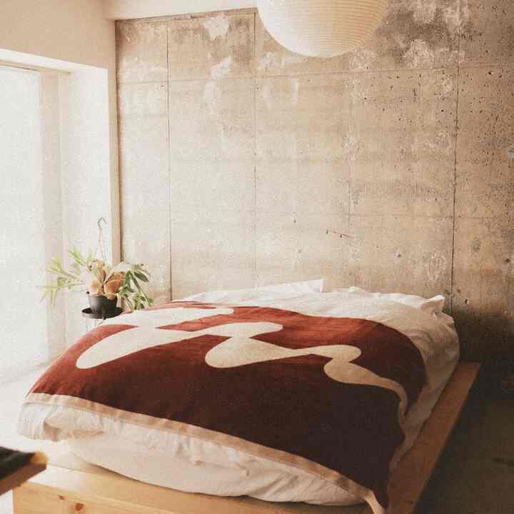 ナチュラルなトーンの寝室、ワインレッドのブランケットとコンクリート壁が調和した温かみのある空間