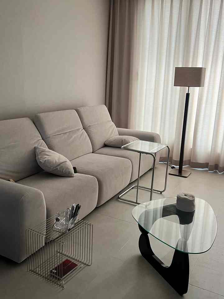 明るいベージュトーンのリビング、ソファと透明なガラステーブル、シルバーのサイドテーブル、フロアランプが配置されたモダンでミニマルな空間