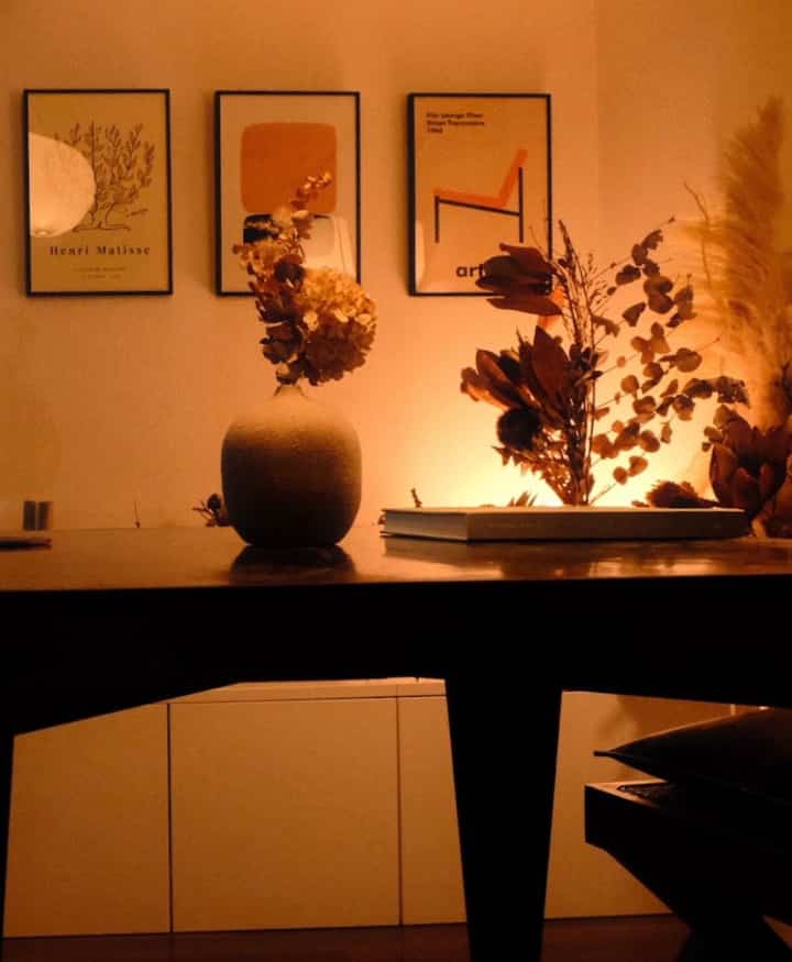 温かみのあるオレンジ色調の1K空間、中央に花瓶とドライフラワー、壁に3つのポスターが配置された居心地のよいフレンチカントリースタイルのインテリア