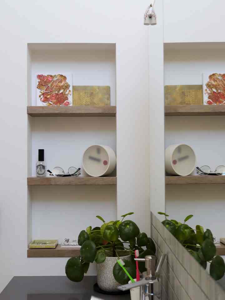 白い壁とナチュラルな木製棚に時計、額縁、植物が配置された洗面台横のモダンなバスルーム