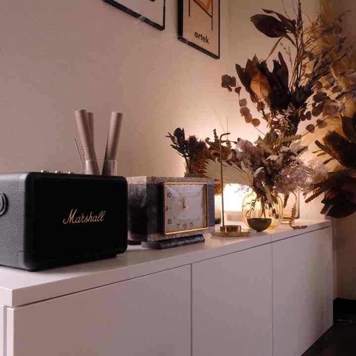 ホワイトの収納家具の上にマシュースピーカーと時計、ゴールドの香立て、ドライフラワーが配置されたナチュラルトーンの空間