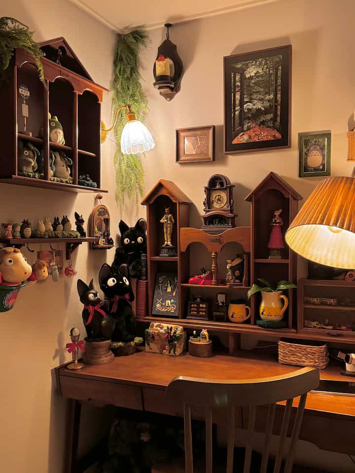 温かみのある照明が灯る寝室の一角、木製の机と椅子、ジブリキャラクターのコレクションが配置された印象的な空間
