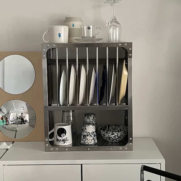 White-walled kitchen space featuring a stainless steel shelf with neatly arranged plates and cups, showcasing a minimal aesthetic