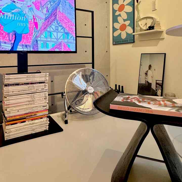 グレーのコンクリート壁とホワイトテーブルのあるモダンな一人暮らしリビング、シルバーメタリックの扇風機と壁のポスターが配置された清潔感のある空間