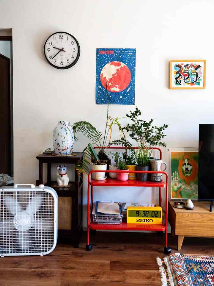 白い壁と木目調の床のリビング、赤い収納カートに植物が置かれ、壁には壁時計とポスターが飾られた雰囲気のある空間