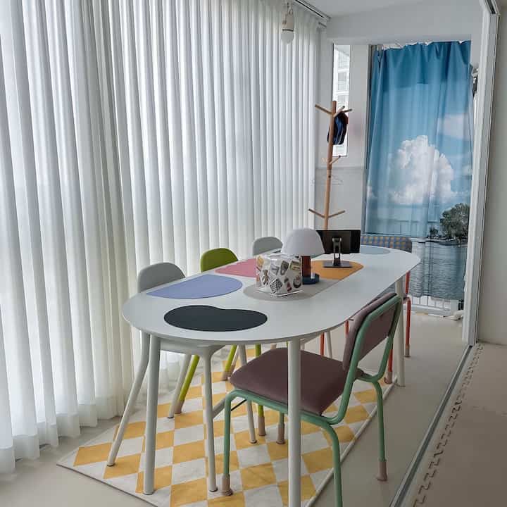 Bright dining room in white and natural tones featuring an oval white dining table with multicolored chairs, coat rack, and balcony curtains creating a cozy atmosphere