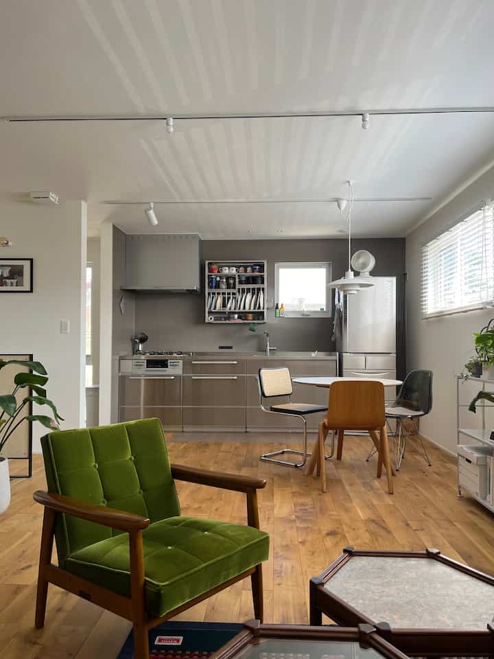 木目調の床にミッドセンチュリーのグリーンアームチェア、ステンレスキッチンとダイニングチェアが配置された明るいキッチン兼ダイニングルームの空間