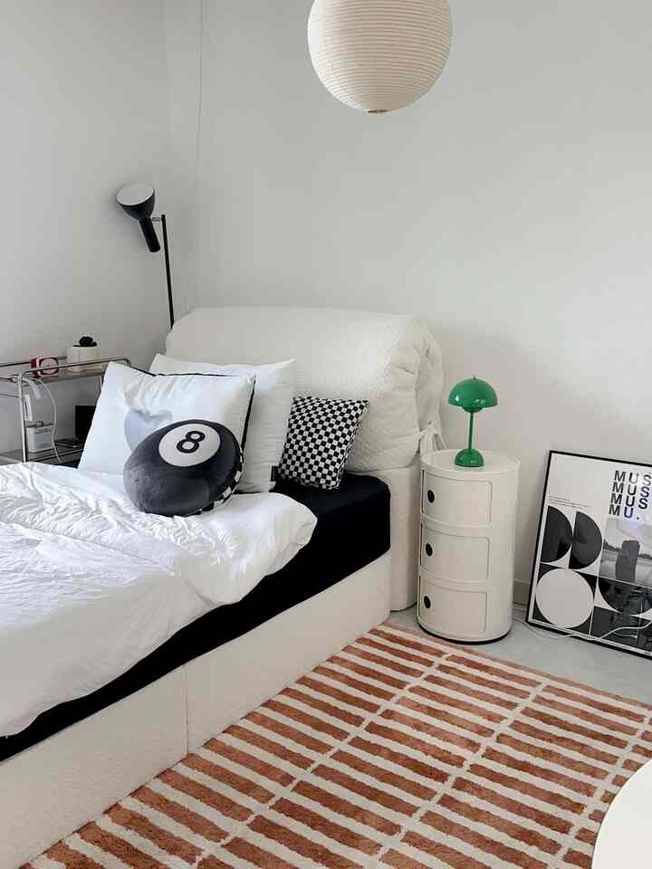 Simple modern bedroom in white and monotone tones featuring a white bed, nightstand, and a brown striped rug