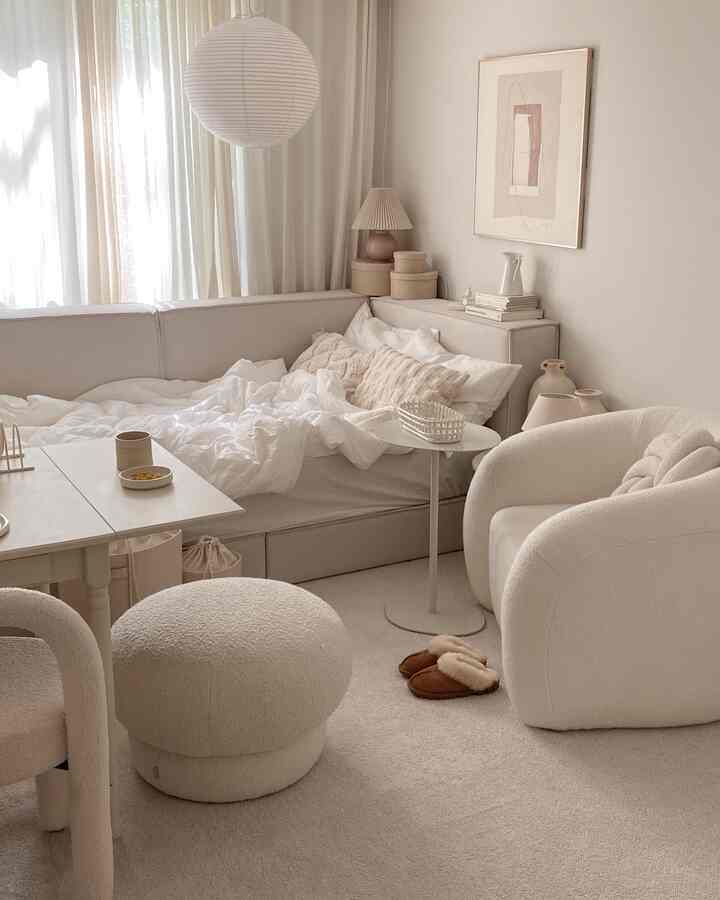 ナチュラルな色調のワンルーム寝室、ベージュのソファとラグ、曲線的なアームチェアとスツールが配された雰囲気のある空間