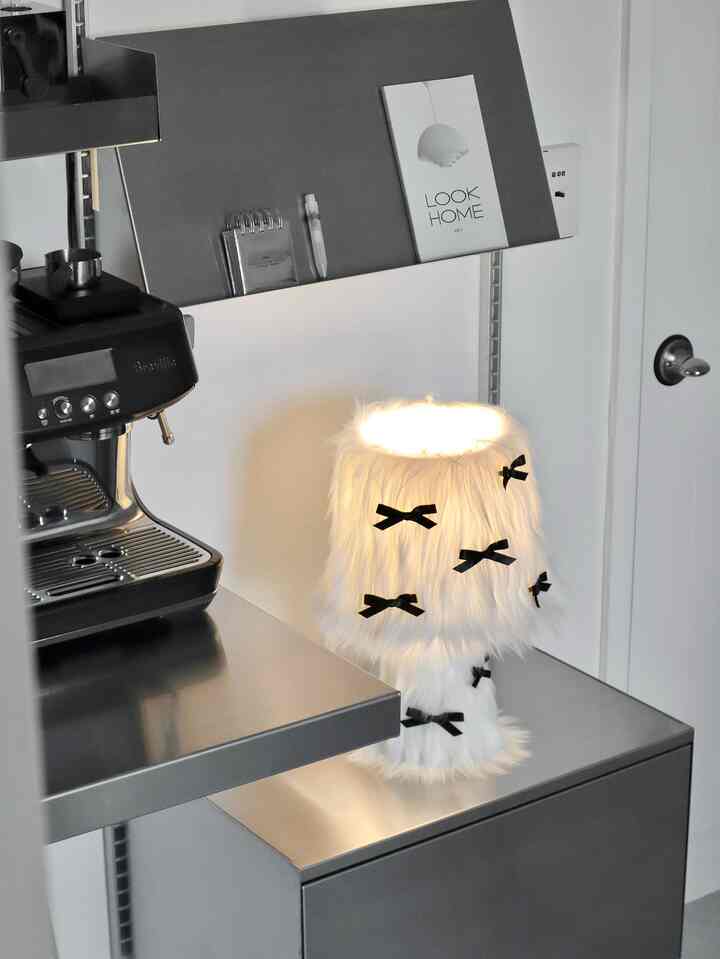 白とシルバーのトーンが印象的なキッチン、柔らかな毛並みのテーブルランプと黒いエスプレッソマシンが配置された雰囲気のあるインテリア